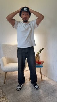 Person trägt eine schwarze Kappe mit der Aufschrift "FORMER MERCH" in Silber, weißes T-Shirt, dunkle Jeans und schwarz-weiße Sneaker, steht drinnen.