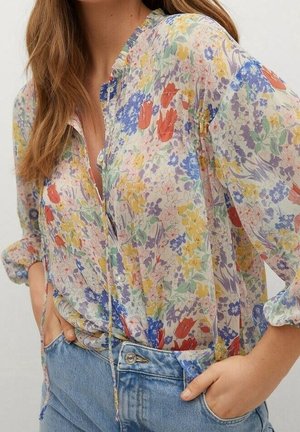 Femme portant un chemisier transparent à fleurs rouges, jaunes, bleues et violettes, rentrée dans un jean bleu clair, les mains dans les poches.
