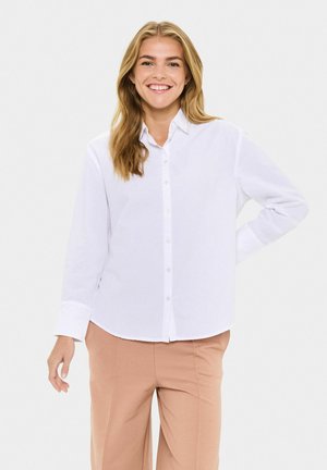 Young woman smiling with loose blonde hair wearing a white button-up shirt and beige pants, standing against a plain background.
