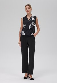 Blusa nera senza maniche con motivo floreale bianco, abbinata a pantaloni neri sartoriali e tacchi neri. Tessuto liscio, design aderente.