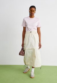 Ljusrosa bomullst-shirt i kombination med en krämfärgad maxikjol med elastisk midja och cargofickor, bärs med grå sneakers och en liten brun väska.