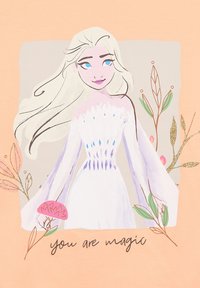 Koszulka w kolorze brzoskwini, z ilustracją postaci o długich białych włosach, ubraną w białą sukienkę, otoczoną kwiecistymi akcentami i tekstem "jesteś magią."