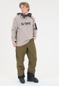 Lys beige hættetrøje med sort "SLOPE" logo, parret med olivengrønne bukser med sidelommer og sorte detaljer ved manchetterne.