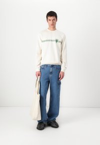 White crewneck sweatshirt with green stripes and logo, paired with loose blue denim jeans and black textured sneakers. Beige tote bag held in hand.
