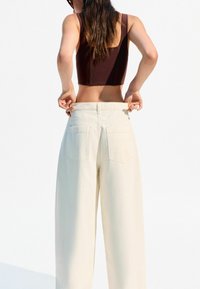 Brown cropped tank top paired with high-waisted white wide-leg trousers, featuring back pockets and a relaxed fit. Simple, minimalist design.