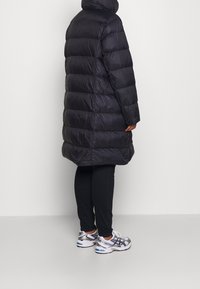 Schwarze Pufferjacke mit hohem Kragen, horizontalem Steppmuster, langen Ärmeln und knielangem Schnitt. Getragen mit schwarzen Hosen und Turnschuhen.