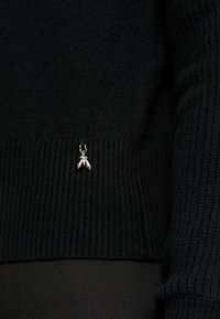 Černý žebrovaný pletený svetr s malou stříbrnou kovovou broží ve tvaru hmyzu připevněnou blízko spodního lemu.