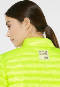 Neon green lightweight jacket with quilted texture, high collar, and a label on the back reading "SENSES .THE LABEL" with minimalist design details.