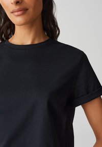 T-shirt nera in cotone con scollo rotondo, maniche corte arrotolate e texture liscia. Il tessuto sembra leggero e informale.