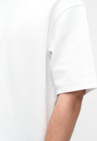 Nærbilde av en persons arm iført en enkel hvit kortermet skjorte på en ensfarget hvit bakgrunn.