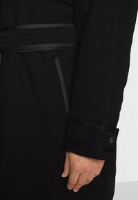 Svart kappa med slät tyg, vertikala ränder vid midjan och ärmsluten, samt en knappaccent på ärmsluten. Sidoficka synlig.