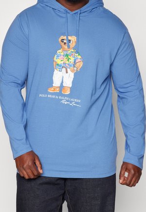 Blauer Hoodie mit einem Bären, der eine Sonnenbrille, ein buntes Hawaiihemd, weiße Hose und orangefarbene Schuhe trägt mit dem Schriftzug „Polo Bear by Ralph Lauren“.