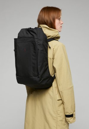 Schwarzer Rucksack mit rechteckiger Form und glatter Oberfläche, ausgestattet mit einer Reißverschlusstasche, getragen von einer Person, die einen gelben Mantel anhat.