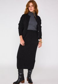 Cardigan noir avec des paillettes dorées, col roulé gris, jupe midi noire. Bottes noires montantes avec des talons carrés. Tissu texturé, design ajusté.