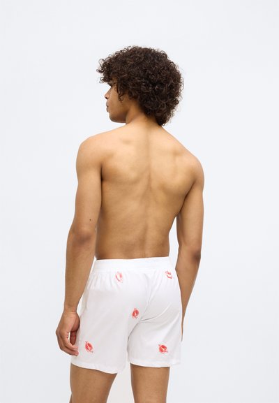 Pantaloni scurți de baie albi cu broderie florală roșie, talie elastică și croială lejeră. Prezintă o textură netedă și o lungime scurtă.