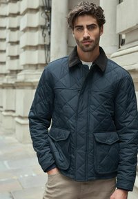Navy gesteppten Jacke mit einem dunklen Kragen, zwei vorderen Taschen und einem Reißverschluss. Über einem hellen Pullover getragen, mit tailliertem Schnitt.