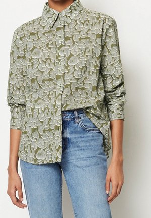 Persoon die een olijfgroen hemd met lange mouwen draagt met een wit bladpatroon en lichtblauwe spijkerbroek, staand tegen een effen muur.