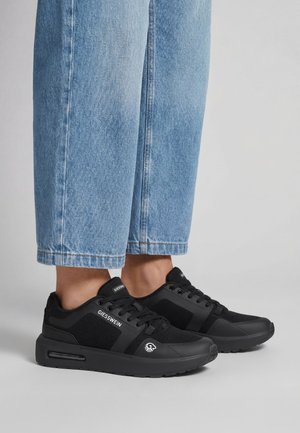 Beine in hellblauen Jeans und schwarzen Giesswein-Sneakern mit Mesh-Einsätzen und dicken Sohlen, stehend auf einer grauen Oberfläche.