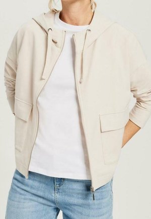 Kvinde iført lysebeige hættetrøje med lynlås og forlommer over en hvid t-shirt og lyseblå jeans, hænderne bag ryggen.