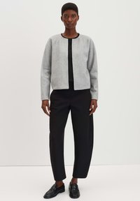 Grijze textuur jacket met een ronde halslijn, gecombineerd met een zwarte top en losse zwarte broek. Zwarte nette schoenen maken de outfit compleet.