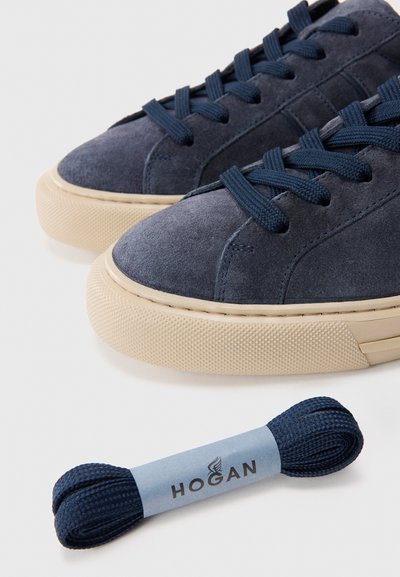 Pair of navy blue suede sneakers with beige rubber soles and matching navy blue spare shoelaces labeled Hogan on a white surface.