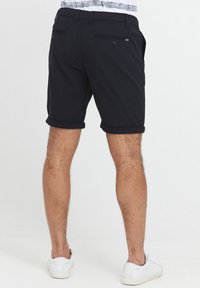 Sorte rullede shorts laget av stoff, med to baklommer med knapper. Personen har på seg hvite joggesko.