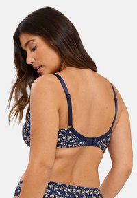 Sujetador de color azul marino con estampado floral, con tirantes ajustables, cierre de gancho y ojo, y una textura suave que muestra pequeñas flores amarillas y blancas.