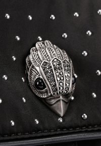 Czarna tkaninowa torba z metaliczną, srebrną broszką w kształcie ptaka, ozdobioną czarnymi kamieniami i otoczoną małymi srebrnymi ćwiekami.