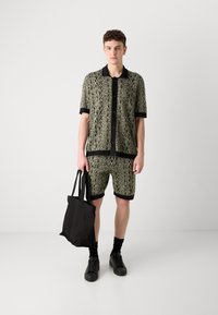 Black and green patterned shorts and button-up shirt set, made of lightweight fabric. Model holds a black tote bag, wearing black shoes and socks.