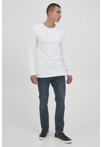 Langermet hvit henley-skjorte med tre knapper ved kragen, paret med mørkeblå jeans og svarte joggesko. Jevn tekstur og tettsittende design.