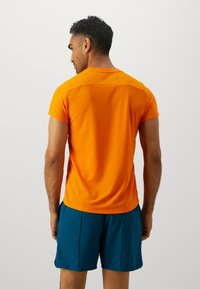 Orange trænings t-shirt med korte ærmer, glat tekstur og lige pasform. Bæres med blå shorts, der viser et enkelt og funktionelt design.