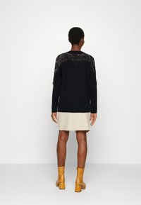 Schwarzer Pullover mit Spitzenakzenten an den Schultern, kombiniert mit einem beigen Rock und gelben Knöchelstiefeln. Glatte Textur, lässiges Design, figurbetonter Stil.