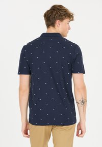 Mörkblå polotröja i bomull, med ett subtilt vitt mönster. Korta ärmar och klassisk krage. Bakifrån syns en tatuering på armen.