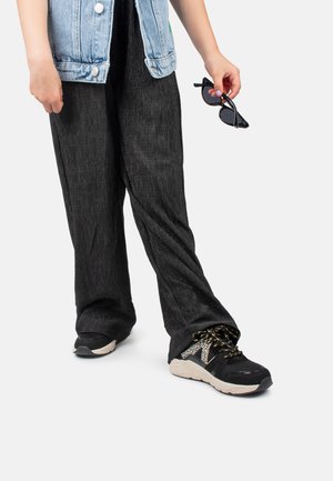 Gilet en jean bleu clair, pantalon large noir texturé et baskets noires avec accents en mesh ; lunettes de soleil tenues à la main.
