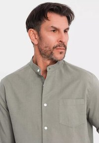 Camicetta verde chiaro a maniche lunghe, con colletto e tasca. Presenta bottoni bianchi e un tessuto liscio e texturizzato.