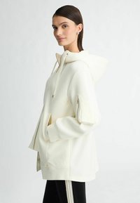 Crème hoodie met een rits aan de voorkant en zijzakken, voorzien van een ritszak op de mouw en een relaxte pasvorm. Zachte stofstructuur.