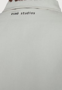 Achterkant van een lichtgrijs overhemd met kraag, met "esmé studios" in het zwart geborduurd vlak bij de halslijn.