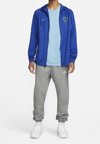 Blå zip-jakke med hætte, lyseblå t-shirt under, grå sweatpants og hvide sportsko, med et logo på jakken.