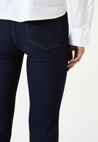 Jean en denim bleu foncé à coupe ajustée, avec deux poches arrière ornées d'un motif de couture discret. Associé à une chemise blanche boutonnée.