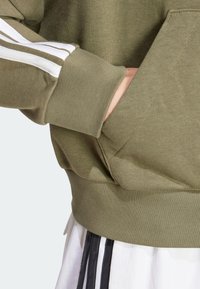 Grön sweatshirt med vita ränder på ärmarna, ribbade muddar och midja, samt en ficka på framsidan för funktionalitet.