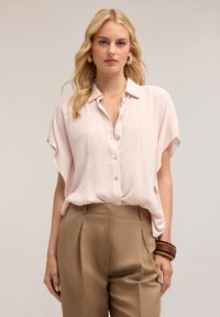 Blusa button-up rosa chiaro con maniche corte, tessuto morbido, vestibilità ampia. Abbinata a pantaloni a vita alta color beige, con pince e un bracciale in legno.