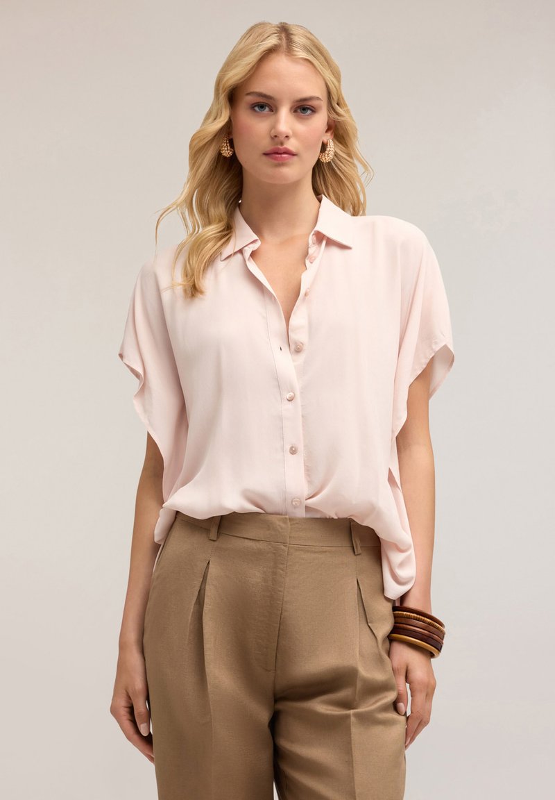 Blusa button-up rosa chiaro con maniche corte, tessuto morbido, vestibilità ampia. Abbinata a pantaloni a vita alta color beige, con pince e un bracciale in legno.