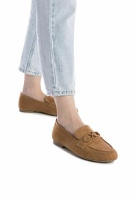 Mocasines de ante marrón con suela plana, que presentan un detalle decorativo de lazo en la puntera. Llevados con jeans de tiro bajo en un tono azul claro.