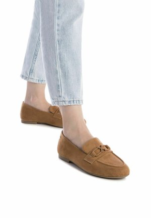 Mocasines de ante marrón con suela plana, que presentan un detalle decorativo de lazo en la puntera. Llevados con jeans de tiro bajo en un tono azul claro.