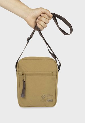 Bolso bandolera de tela marrón con correa ajustable, bolsillo frontal con cremallera y detalle de logo impreso en la parte inferior derecha. Diseño duradero y compacto.