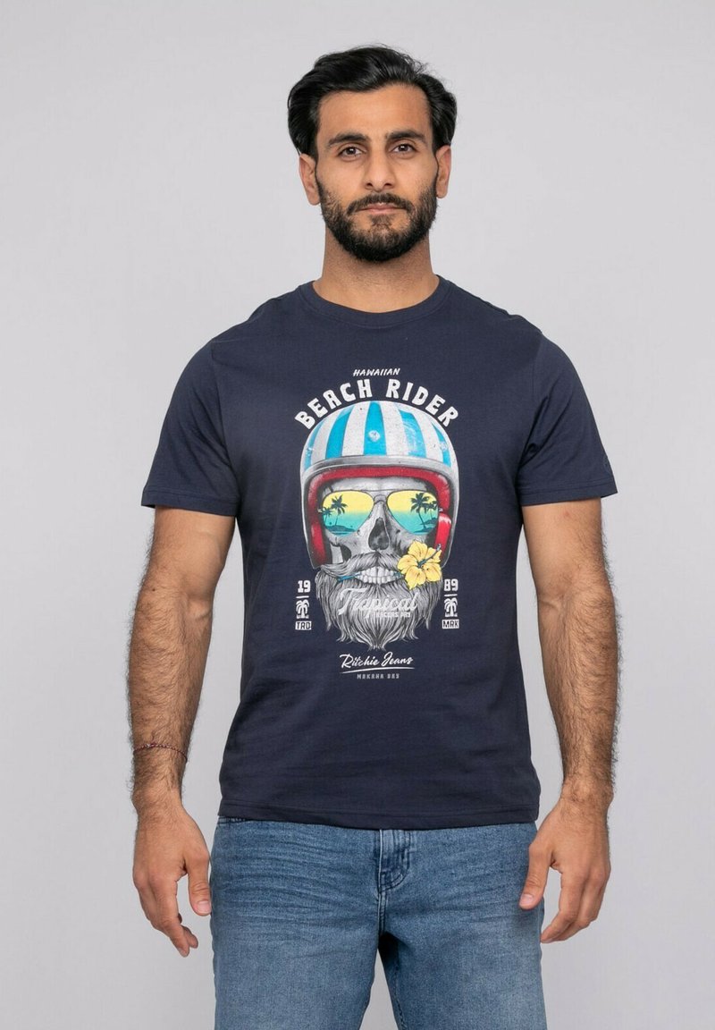 Homme barbu portant un t-shirt bleu marine avec un graphisme coloré de crâne arborant un casque, des lunettes de soleil reflétant des palmiers, et une fleur jaune.