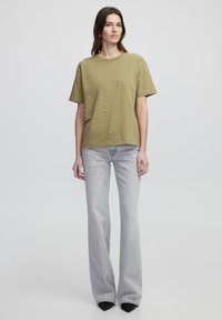 Camiseta verde oliva de manga corta, con pequeños adornos blancos dispersos; combinada con pantalones de campana gris claro y zapatos negros.