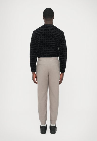 Μαύρο πουλόβερ με υφή και γεωμετρικό μοτίβο, ανοιχτό γκρι παντελόνι με στενή γραμμή και διακριτική υφή, και μαύρα αθλητικά παπούτσια. Πλάγια όψη από πίσω, ουδέτερο φόντο.