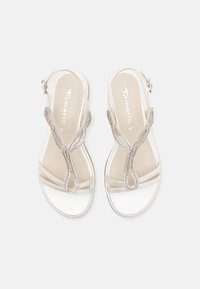 Beige slingback sandals with a textured strap design featuring metallic accents and a smooth, shiny surface. Closed toe with buckle detail.