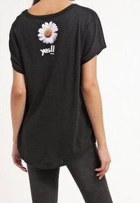 Černé tričko s krátkým rukávem s bílým grafickým motivem sedmikrásky a textem "ano!!" na zadní straně; měkká tkanina a volný střih.